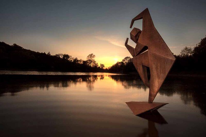 A large, bronze, angular sculpture of a figure stretching in a ballet pose in front of a lake at sunset.