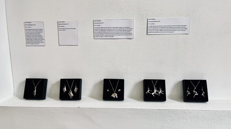 A photograph of five jewellery boxes displaying various pendants of people in motion. These are on display on a gallery shelf. They are accompanied by descriptions of each piece.